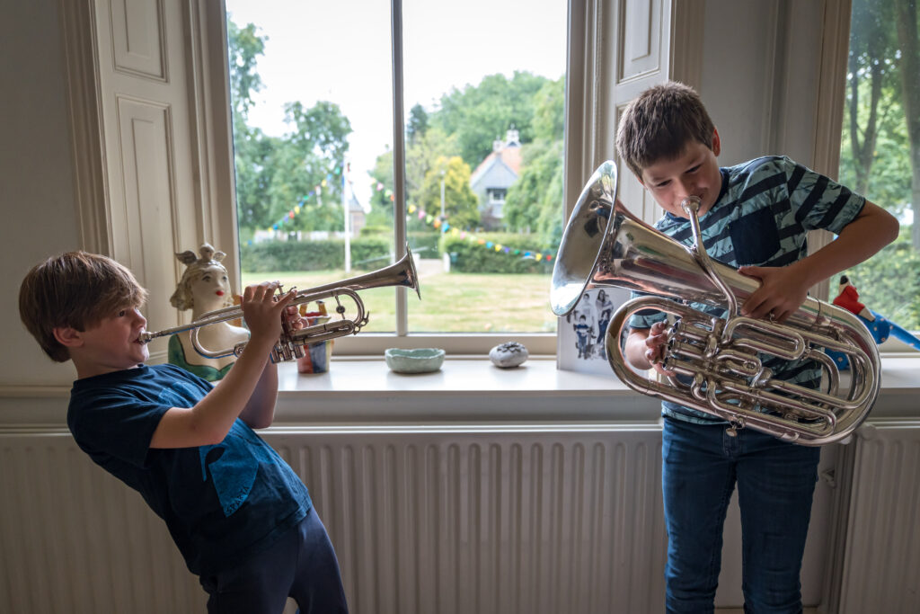 Twee kinderen zijn samen muziek aan het maken voor een raam. De ene speelt trompet, de andere bariton. Op de achtergrond is een grasveld te zien waaar vlaggetjes hangen.
