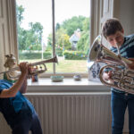Twee kinderen zijn samen muziek aan het maken voor een raam. De ene speelt trompet, de andere bariton. Op de achtergrond is een grasveld te zien waaar vlaggetjes hangen.